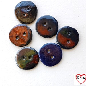 Peut inclure: Un assortiment de boutons en céramique faits à la main de différentes couleurs. Les boutons sont ronds avec deux trous et présentent une finition brillante. Les couleurs incluent le marron foncé, l'orange, le bleu et un mélange de vert et de rouge. Les boutons sont disposés sur une surface blanche.