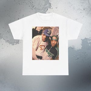 Puede incluir: Una camiseta blanca de manga corta con un estampado gráfico rectangular centrado de dos personas. Una persona lleva una gorra azul y una chaqueta blanca, mientras que la otra lleva una cinta verde y una chaqueta deportiva negra y blanca.