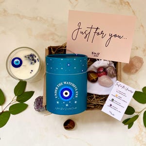 Puede incluir: Un juego de regalo con una vela azul con un dise&ntilde;o de ojo turco, una peque&ntilde;a bolsa de cristales y una tarjeta que dice "Just for you...". La vela est&aacute; en un recipiente cil&iacute;ndrico con el texto "UNDER THE WATCHFUL EYE".