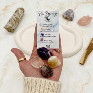 May include: A hand holding a collection of crystals, including clear quartz, citrine, amethyst, and rhodonite. A card with the words "New Beginnings" and crystal properties is also held. A sage bundle and palo santo stick are on the white tray.