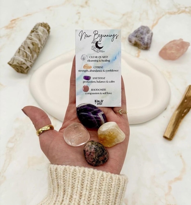May include: A hand holding a collection of crystals, including clear quartz, citrine, amethyst, and rhodonite. A card with the words "New Beginnings" and crystal properties is also held. A sage bundle and palo santo stick are on the white tray.