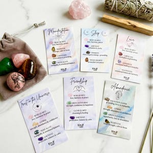 May include: A collection of crystals and affirmation cards. A gray drawstring bag holds green, pink, and brown stones. Cards with watercolor backgrounds list crystal properties. Sage bundles, lavender, and a rose quartz crystal are also present.