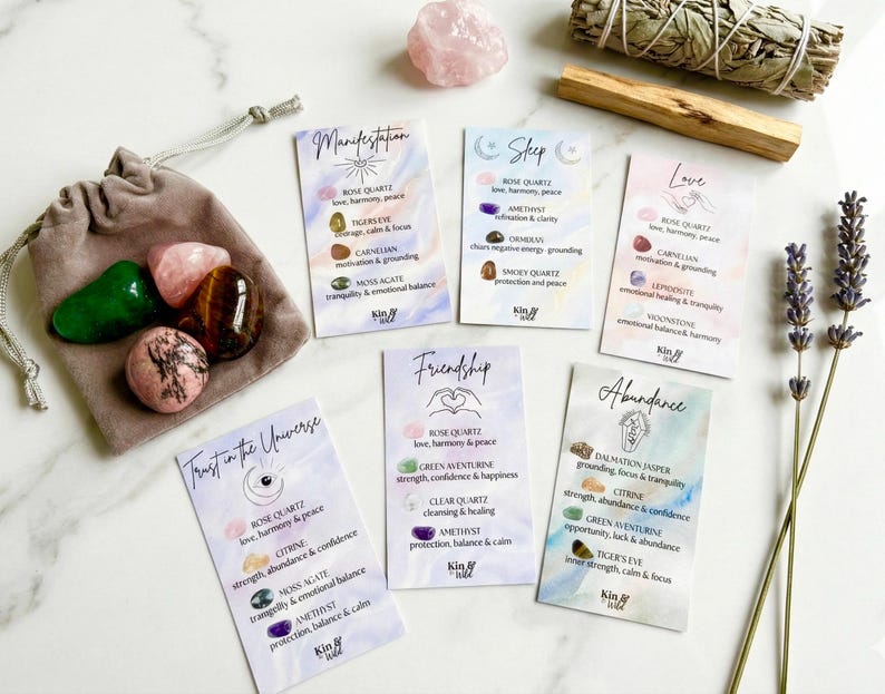 May include: A collection of crystals and affirmation cards. A gray drawstring bag holds green, pink, and brown stones. Cards with watercolor backgrounds list crystal properties. Sage bundles, lavender, and a rose quartz crystal are also present.