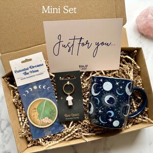 May include: A gift box containing a dark blue mug with a celestial moon and star pattern, a Celestial Dreams The Moon air freshener, a Rose Quartz mushroom keyring, and a card with the text Just for you and Kin & Wild, all nestled in paper.