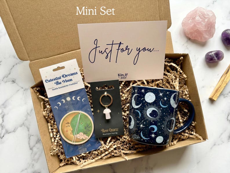 May include: A gift box containing a dark blue mug with a celestial moon and star pattern, a Celestial Dreams The Moon air freshener, a Rose Quartz mushroom keyring, and a card with the text Just for you and Kin & Wild, all nestled in paper.