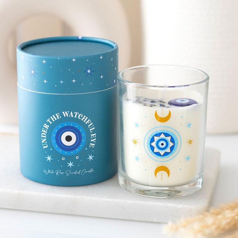Puede incluir: Una vela perfumada con rosas blancas en un vaso transparente con un dise&ntilde;o de ojo azul y dorado. La vela est&aacute; junto a una caja cil&iacute;ndrica azul con el texto "UNDER THE WATCHFUL EYE" y un dise&ntilde;o de ojo a juego.