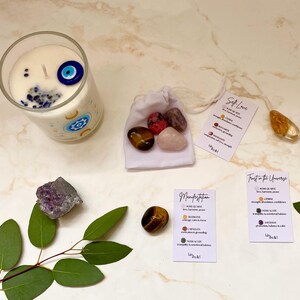 Puede incluir: Una vela en un vaso transparente con un dise&ntilde;o de ojo turco, rodeada de cristales y peque&ntilde;as tarjetas. Las tarjetas tienen texto que dice "Self Love", "Manifestation" y "Trust in the Universe". Una bolsa blanca contiene varias piedras.