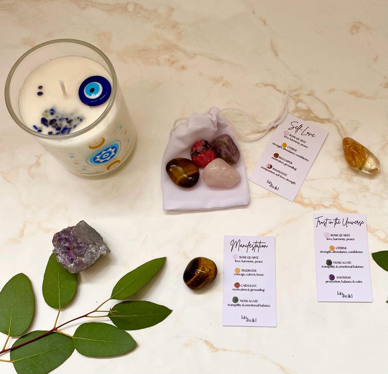 Puede incluir: Una vela en un vaso transparente con un dise&ntilde;o de ojo turco, rodeada de cristales y peque&ntilde;as tarjetas. Las tarjetas tienen texto que dice "Self Love", "Manifestation" y "Trust in the Universe". Una bolsa blanca contiene varias piedras.