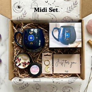 Op de afbeelding: Een geschenkdoos met het label "Midi Set" met een marineblauwe mok met een oog- en maansikkelontwerp, een kaars, een sleutelhanger, een kaart met de tekst "Just for you" en amethiststenen. De doos is gevuld met bruin papier en versierd met hemelse illustraties.