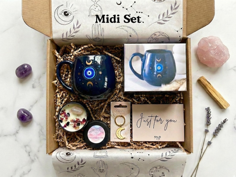 Op de afbeelding: Een geschenkdoos met het label "Midi Set" met een marineblauwe mok met een oog- en maansikkelontwerp, een kaars, een sleutelhanger, een kaart met de tekst "Just for you" en amethiststenen. De doos is gevuld met bruin papier en versierd met hemelse illustraties.