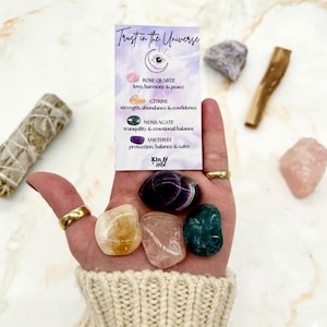 May include: A hand displays a collection of crystals, including rose quartz, citrine, moss agate, and amethyst. A card reads "Trust in the Universe" with crystal properties. Additional items include a sage bundle and a piece of palo santo.