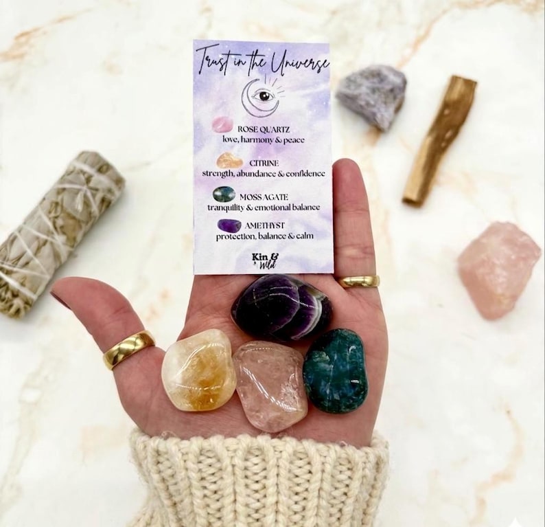 May include: A hand displays a collection of crystals, including rose quartz, citrine, moss agate, and amethyst. A card reads "Trust in the Universe" with crystal properties. Additional items include a sage bundle and a piece of palo santo.