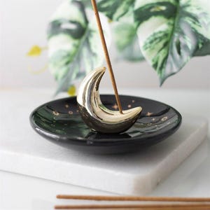May include: A black ceramic incense holder featuring a shiny gold crescent moon in the center. The circular dish is decorated with small gold stars and celestial symbols. A single brown incense stick is inserted into the moon holder.