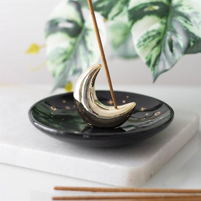 May include: A black ceramic incense holder featuring a shiny gold crescent moon in the center. The circular dish is decorated with small gold stars and celestial symbols. A single brown incense stick is inserted into the moon holder.