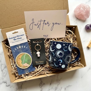 May include: A cardboard gift box containing a mug with a dark blue celestial moon and star pattern, a Celestial Dreams The Moon air freshener, a rose quartz mushroom keychain on a card, and a card with the text Just for you and Kin & Wild.