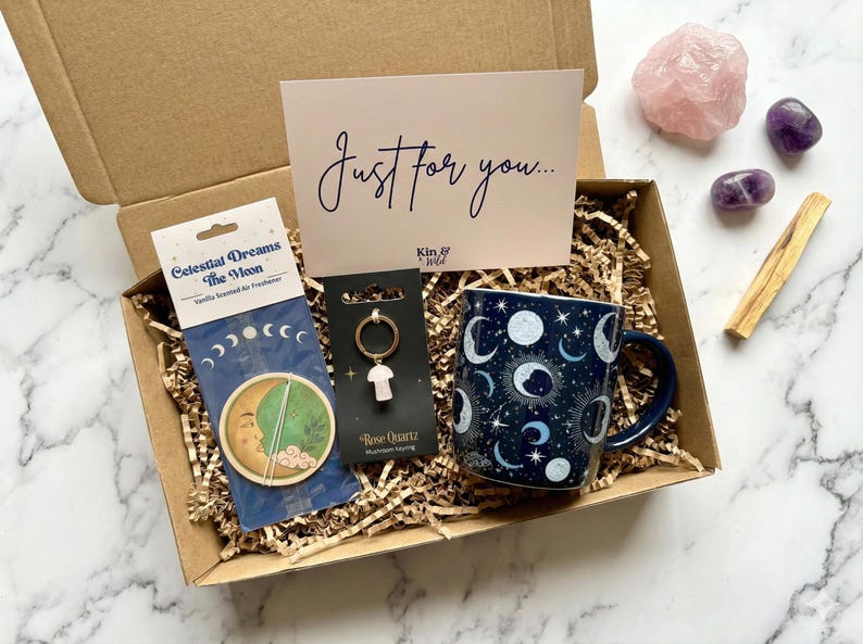 May include: A cardboard gift box containing a mug with a dark blue celestial moon and star pattern, a Celestial Dreams The Moon air freshener, a rose quartz mushroom keychain on a card, and a card with the text Just for you and Kin & Wild.