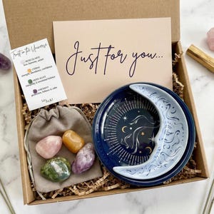 May include: A gift box with a navy blue moon-shaped dish, crystals, a small bag of stones, and a card that says "Just for you...". The box also includes a small card with text and a piece of palo santo.