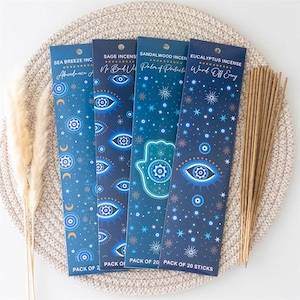 May include: Four vertical rectangular incense packages in shades of blue, each labeled with scents like Sea Breeze, Sage, Sandalwood, and Eucalyptus. The packaging features eye, star, moon, and hamsa hand motifs. A bundle of light brown incense sticks is included.