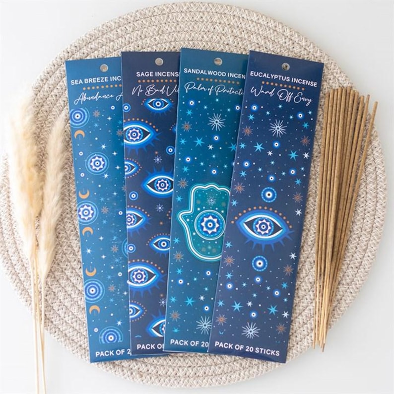 May include: Four vertical rectangular incense packages in shades of blue, each labeled with scents like Sea Breeze, Sage, Sandalwood, and Eucalyptus. The packaging features eye, star, moon, and hamsa hand motifs. A bundle of light brown incense sticks is included.