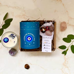 Puede incluir: Un juego de regalo con una vela azul con un dise&ntilde;o de ojo y el texto "UNDER THE WATCHFUL EYE". El juego incluye cristales y una peque&ntilde;a bolsa de piedras. La vela est&aacute; rodeada de hojas de eucalipto y cristales.