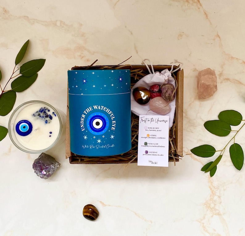 Puede incluir: Un juego de regalo con una vela azul con un dise&ntilde;o de ojo y el texto "UNDER THE WATCHFUL EYE". El juego incluye cristales y una peque&ntilde;a bolsa de piedras. La vela est&aacute; rodeada de hojas de eucalipto y cristales.