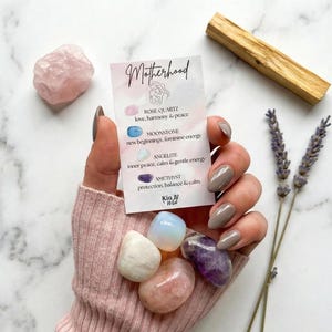 May include: A collection of tumbled gemstones, including rose quartz, amethyst, and moonstone, held in a hand. A card with the word "Motherhood" and gemstone descriptions is also present, along with a palo santo stick and lavender sprigs.