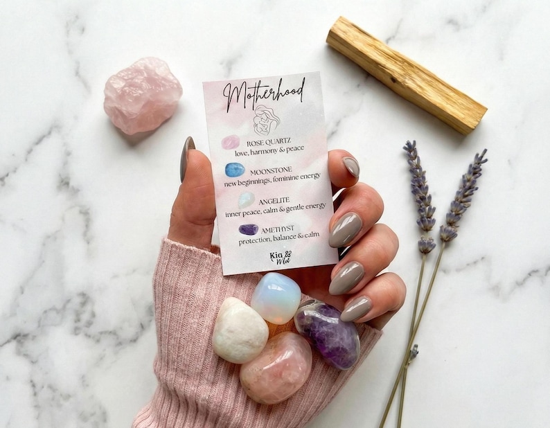 May include: A collection of tumbled gemstones, including rose quartz, amethyst, and moonstone, held in a hand. A card with the word "Motherhood" and gemstone descriptions is also present, along with a palo santo stick and lavender sprigs.