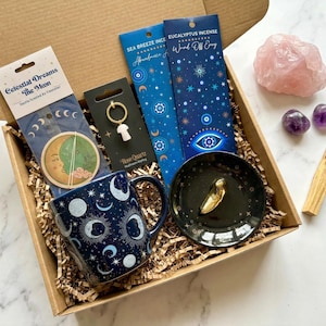 May include: A gift box containing a dark blue mug with moon patterns, a Celestial Dreams air freshener, a rose quartz mushroom keychain, two incense packs, a black trinket dish with a gold charm, a large rose quartz, two amethyst stones, and a palo santo stick.