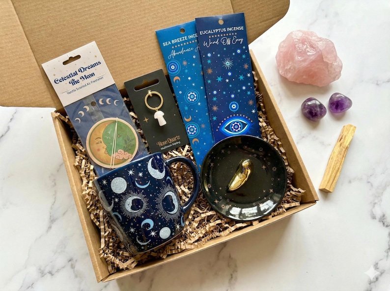 May include: A gift box containing a dark blue mug with moon patterns, a Celestial Dreams air freshener, a rose quartz mushroom keychain, two incense packs, a black trinket dish with a gold charm, a large rose quartz, two amethyst stones, and a palo santo stick.