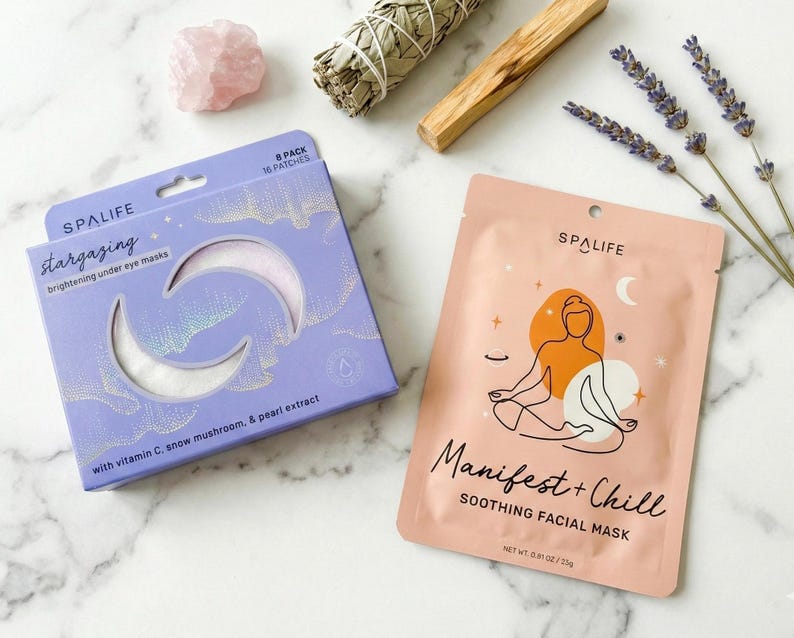 May include: A flat lay featuring spa products. A purple box of "Stargazing" eye masks and a pink "Manifest + Chill" facial mask are displayed. Additional items include a rose quartz crystal, sage bundle, palo santo stick, and lavender sprigs.