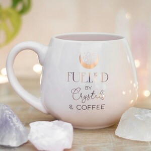 以下が含まれることがあります： 「Fueled by Crystals & Coffee」の金色の文字が入った白いセラミックマグ。マグには三日月とクリスタルのデザインが施されています。アメジストと水晶の結晶がマグの周りに配置されています。
