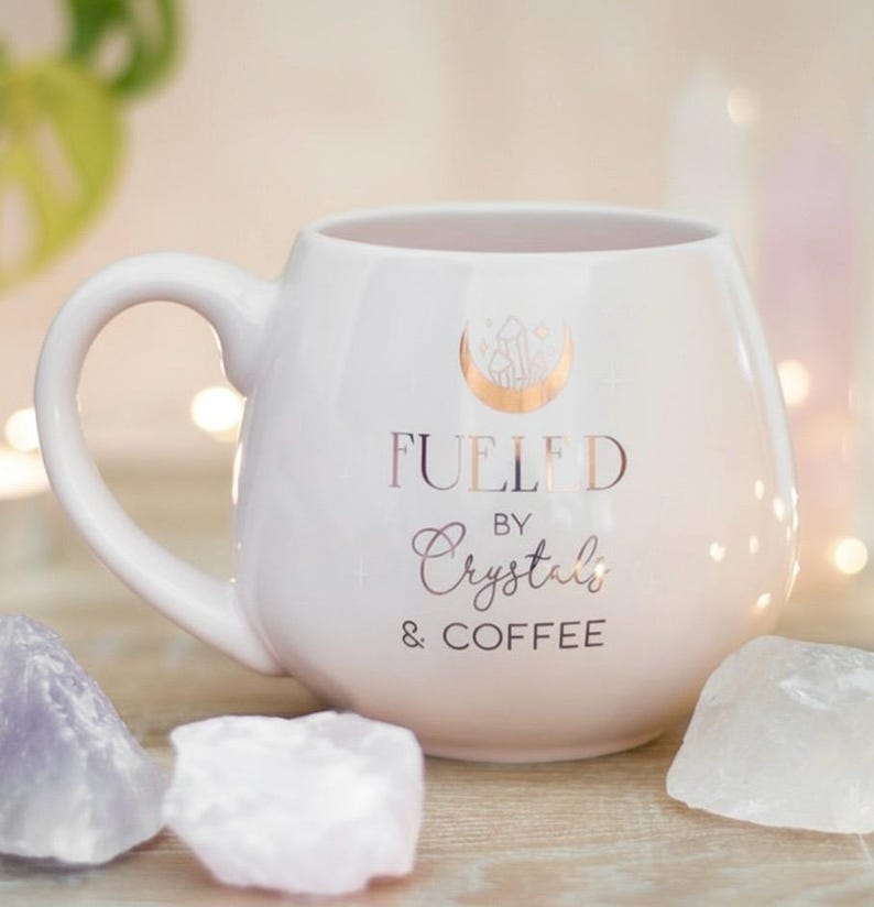 以下が含まれることがあります： 「Fueled by Crystals & Coffee」の金色の文字が入った白いセラミックマグ。マグには三日月とクリスタルのデザインが施されています。アメジストと水晶の結晶がマグの周りに配置されています。