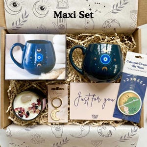 Op de afbeelding: Een geschenkdoos met het label "Maxi Set" bevat een donkerblauwe mok met een oog- en maansikkelontwerp, een kaars, een sleutelhanger, een kaart met de tekst "Just for you" en een zakje. De doos is versierd met hemelse illustraties.