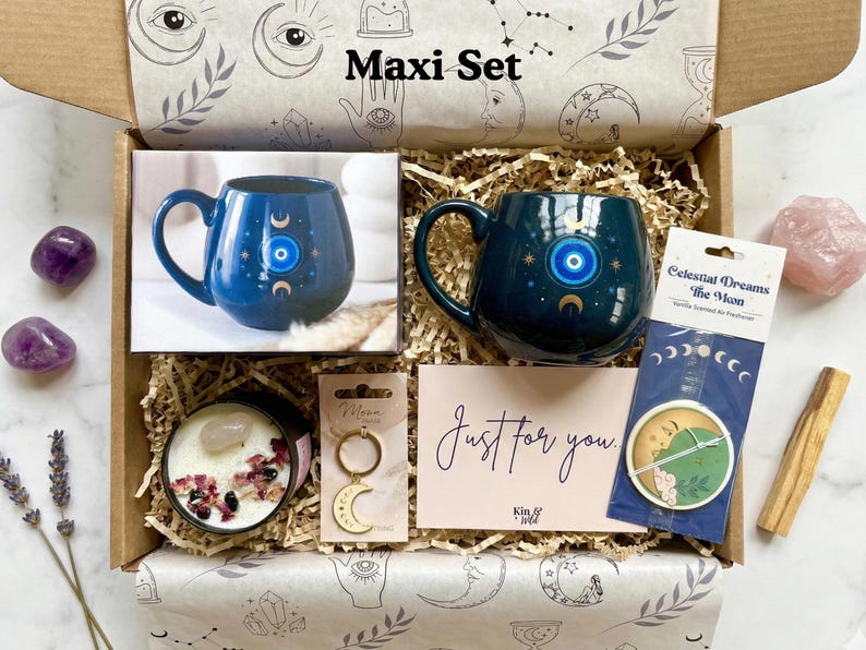 Op de afbeelding: Een geschenkdoos met het label "Maxi Set" bevat een donkerblauwe mok met een oog- en maansikkelontwerp, een kaars, een sleutelhanger, een kaart met de tekst "Just for you" en een zakje. De doos is versierd met hemelse illustraties.