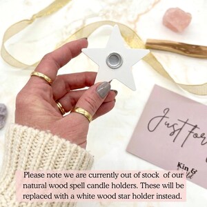 May include: A white star-shaped candle holder with a silver magnetic center is held in a hand. The hand is wearing gold rings and a cream-colored sweater. A pink card with the words "Just For" is in the background.