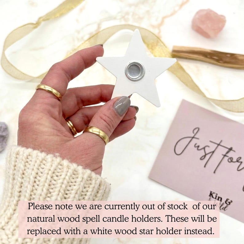 May include: A white star-shaped candle holder with a silver magnetic center is held in a hand. The hand is wearing gold rings and a cream-colored sweater. A pink card with the words "Just For" is in the background.