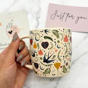 May include: Cream-colored ceramic mug with a whimsical pattern of flowers, hearts, and swallows. The mug has a curved handle and is held by a hand. A card with the text "Just for you..." is in the background.