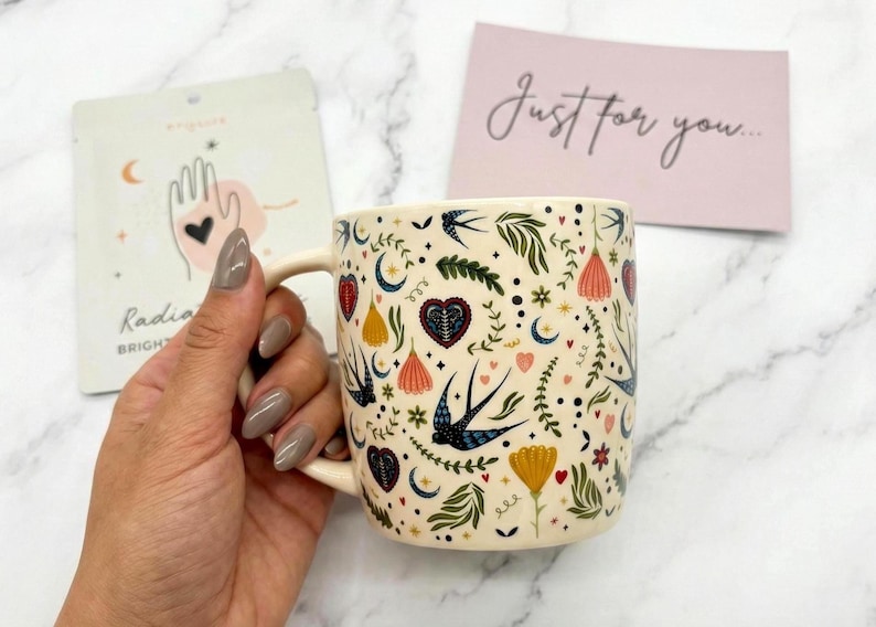 May include: Cream-colored ceramic mug with a whimsical pattern of flowers, hearts, and swallows. The mug has a curved handle and is held by a hand. A card with the text "Just for you..." is in the background.