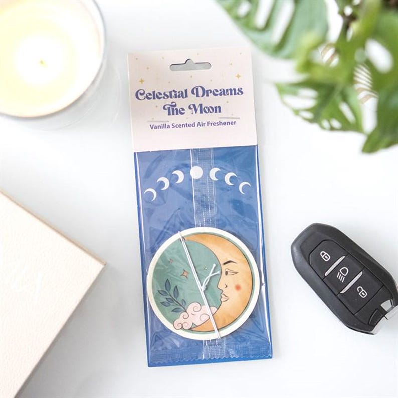 May include: A Celestial Dreams The Moon vanilla scented air freshener in clear plastic packaging. The circular freshener features a crescent moon face with a green side, a leafy branch, and pink clouds. The header displays the product name.