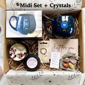 Op de afbeelding: Een geschenkdoos met de tekst "Midi Set + Crystals" bevat een blauwe mok met een oogontwerp, een kaars, kristallen, een sleutelhanger en een kaart met de tekst "Just for you...". De doos is gevuld met decoratief papier en andere items.
