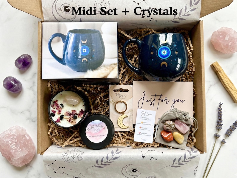 Op de afbeelding: Een geschenkdoos met de tekst "Midi Set + Crystals" bevat een blauwe mok met een oogontwerp, een kaars, kristallen, een sleutelhanger en een kaart met de tekst "Just for you...". De doos is gevuld met decoratief papier en andere items.