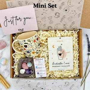 May include: A gift box labeled "Mini Set" contains a floral-decorated mug, a face mask, crystals, and a card that says "Just for you...". The box is filled with shredded paper and includes a bundle of lavender.