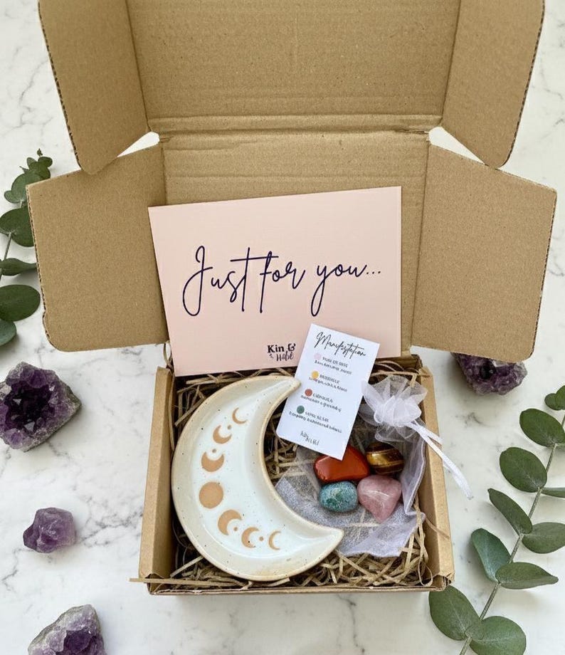 May include: A gift box containing a crescent moon-shaped ceramic dish, crystals, and a card with the text "Just for you...". The dish has a cream base with brown moon phase designs. The box contains a variety of colourful crystals.