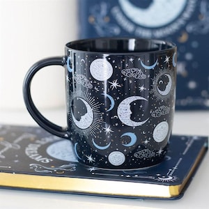 May include: A black ceramic mug featuring a celestial pattern of white and light blue moons, suns, stars, and clouds. The design includes detailed illustrations of crescent moons with faces, full moons, and radiating suns against a dark background.