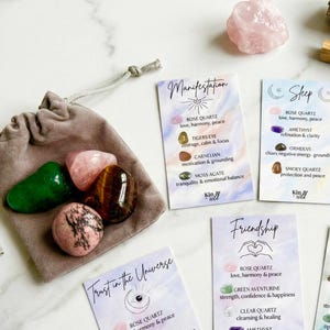 May include: A collection of polished gemstones in a gray velvet pouch, including rose quartz, tiger's eye, and green aventurine. Several affirmation cards with handwritten text are scattered around the pouch, promoting themes of manifestation and friendship.