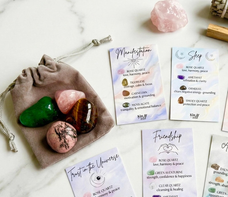 May include: A collection of polished gemstones in a gray velvet pouch, including rose quartz, tiger's eye, and green aventurine. Several affirmation cards with handwritten text are scattered around the pouch, promoting themes of manifestation and friendship.