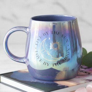 May include: Iridescent ceramic mug with a curved handle. The mug has a blue interior and a pearlescent exterior that shifts colours. The front of the mug features a crescent moon, sun, and the text "LIVE BY THE SUN + LOVE BY THE MOON."