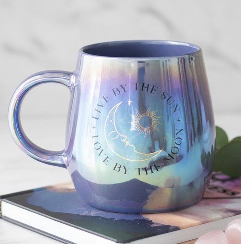 May include: Iridescent ceramic mug with a curved handle. The mug has a blue interior and a pearlescent exterior that shifts colours. The front of the mug features a crescent moon, sun, and the text "LIVE BY THE SUN + LOVE BY THE MOON."