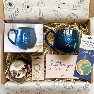 Op de afbeelding: Een open cadeauverpakking met items met een hemels thema. Inclusief een blauwe mok met een oog- en maanontwerp, een kaars, een sleutelhanger, een kaart met "Just for you", een auto-luchtverfrisser, amethiststenen en een rozenkwartskristal.