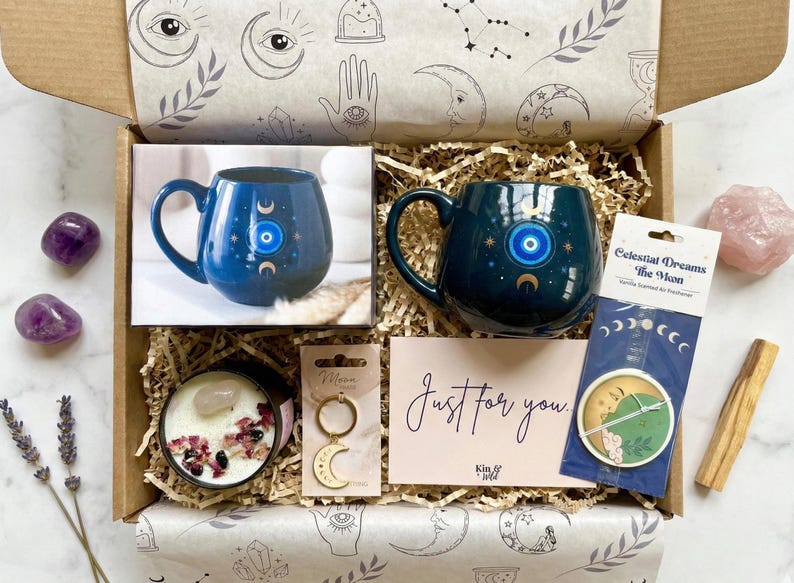 Op de afbeelding: Een open cadeauverpakking met items met een hemels thema. Inclusief een blauwe mok met een oog- en maanontwerp, een kaars, een sleutelhanger, een kaart met "Just for you", een auto-luchtverfrisser, amethiststenen en een rozenkwartskristal.